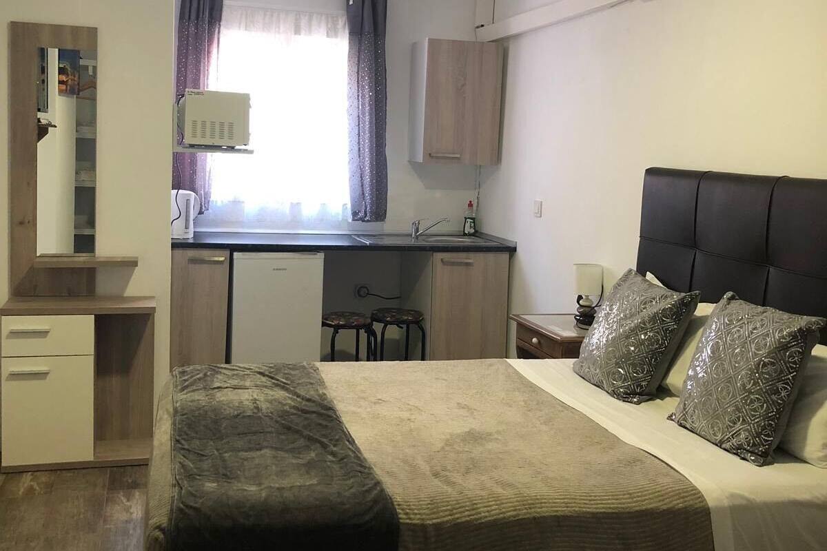 Une chambre à coucher de taille réduite, comprenant un lit, des tables de chevet, une petite kitchenette et une fenêtre avec des rideaux.