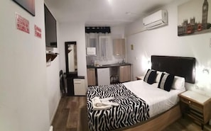 A bedroom with a zebra-patterned bedspread, a wooden nightstand, a desk with a chair, a kitchenette, and an air conditioning unit on the wall.