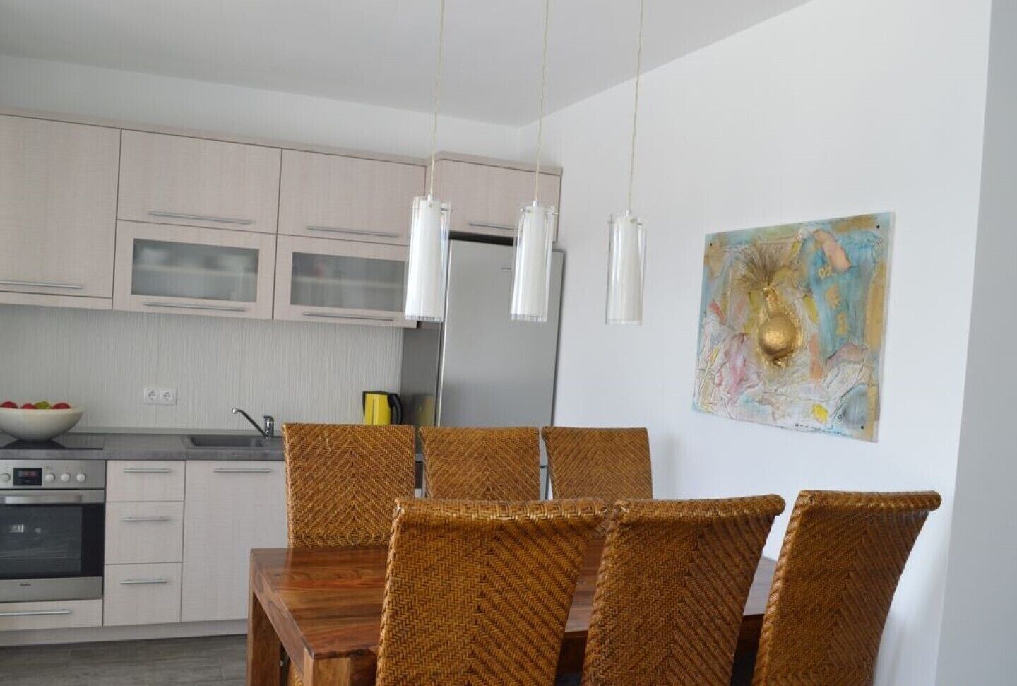 A modern kitchen with white cabinets, a dining table with wicker chairs, and pendant lights.