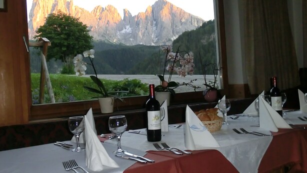 A dining table set with wine glasses, forks, and a bottle of wine, with a mountain view through a window. - Albergo Somont (Ortisei)