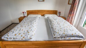 A double bed with floral bedding, a wooden headboard, bedside tables with lamps, a framed picture on the wall, and a window with curtains. - Albergo Somont (Ortisei)