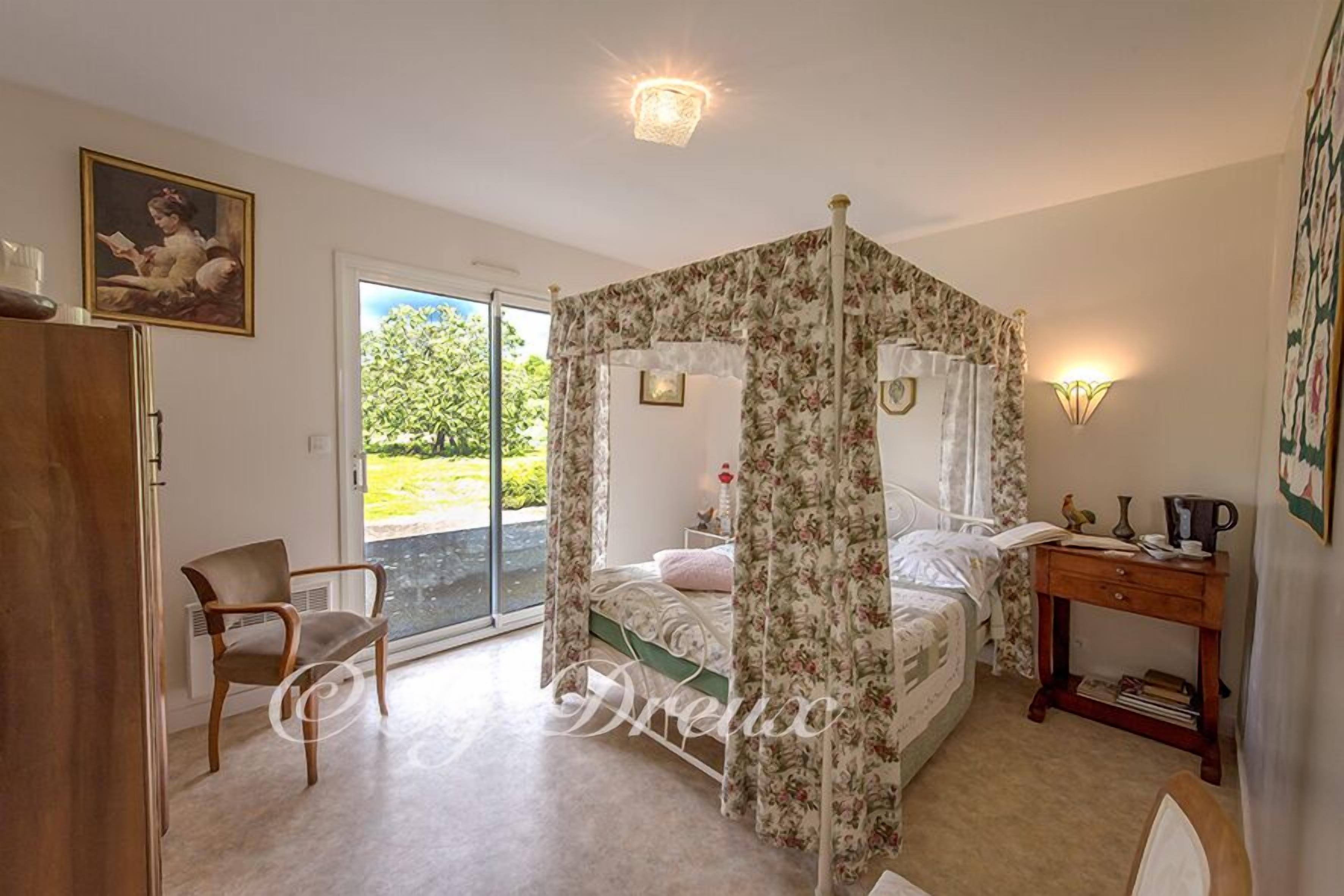 A bedroom with a four-poster bed, a chair, a bedside table, and a view of greenery outside.