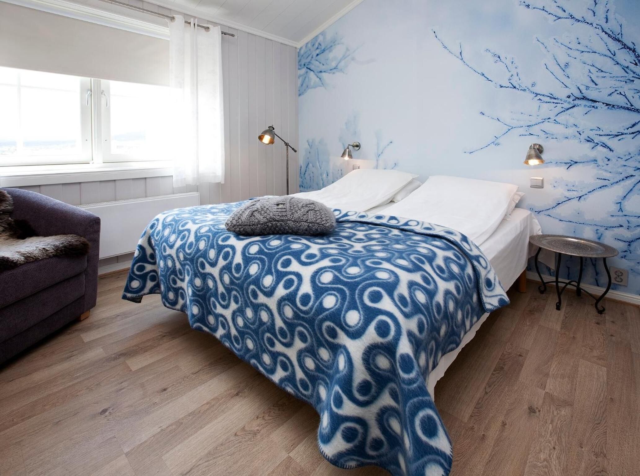 A bedroom with a bed, two bedside lamps, a window with curtains, a chair, and a wall with a tree branch design.