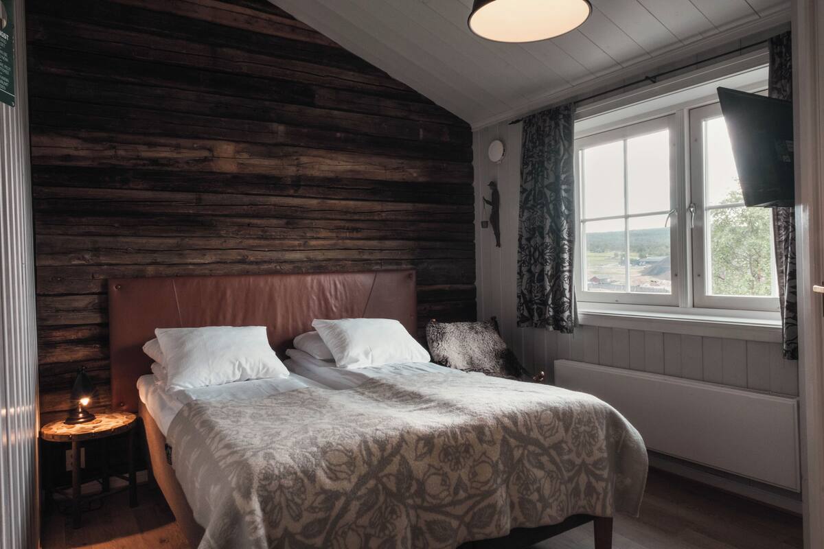 A bedroom with a wooden log wall, a large bed with white bedding, a bedside table with a lamp, and a window with a view of the outdoors.