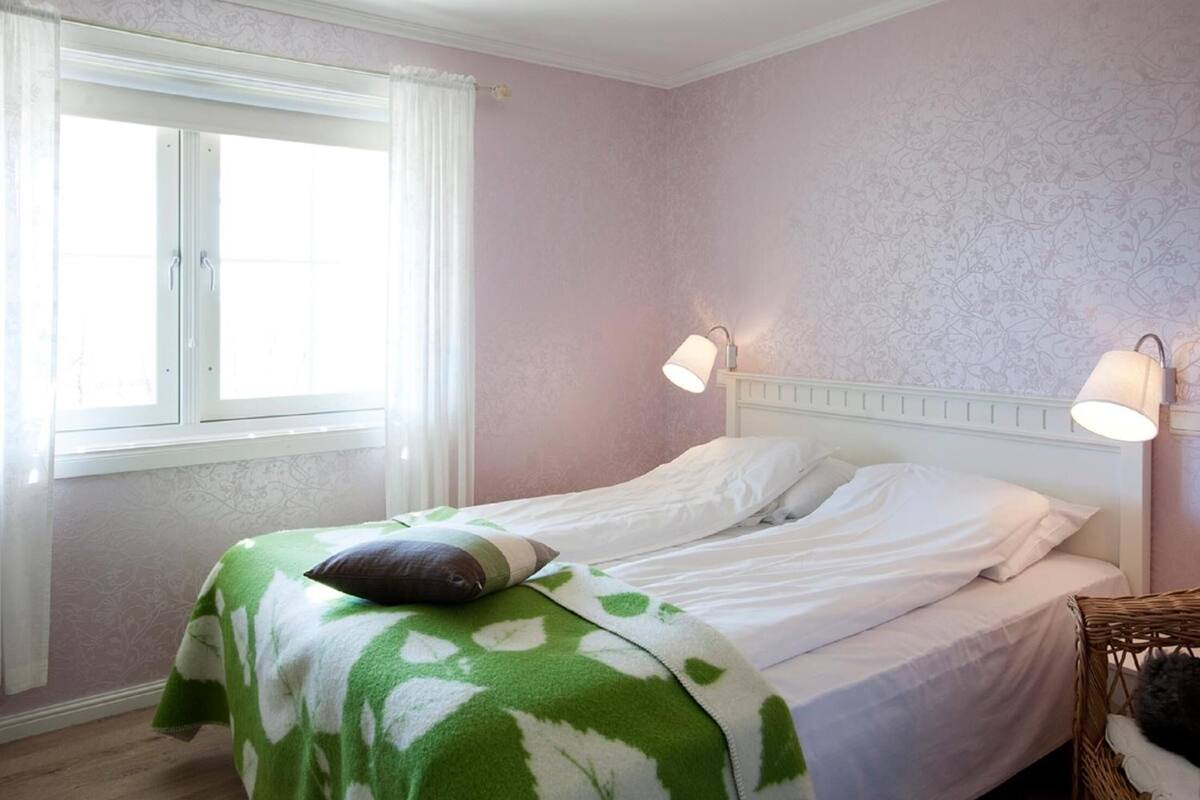 A neatly made bed with a green and white blanket, two bedside lamps, and a window with white curtains.