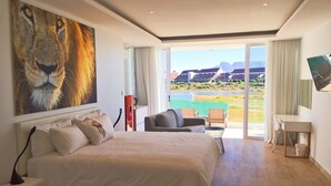 A bedroom with a large bed, a desk, and a chair, a painting of a lion, and a view of a pool and buildings. - Peach on Beach Boutique Guesthouse (Cape Town)