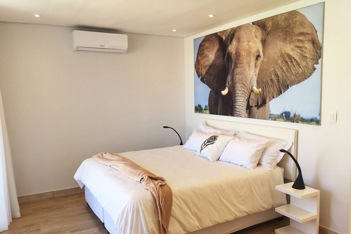 Un dormitorio con cama, aire acondicionado de pared, un cuadro grande de un elefante y una mesita de noche con lámpara.