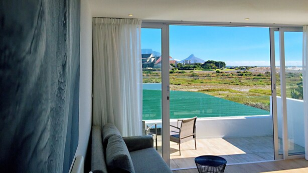 A modern living room with a large window, a sofa, and a view of a green field and distant mountains. - Peach on Beach Boutique Guesthouse (Cape Town)