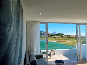 A modern living room with a large window, a sofa, and a view of a green field and distant mountains. - Peach on Beach Boutique Guesthouse (Cape Town)