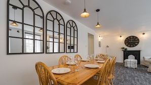 Un coin repas avec une table en bois dressée pour six personnes, entourée de chaises en bois. Trois grands miroirs en forme d’arc ornent le mur, et une cheminée se trouve en dessous, avec une horloge au-dessus.
