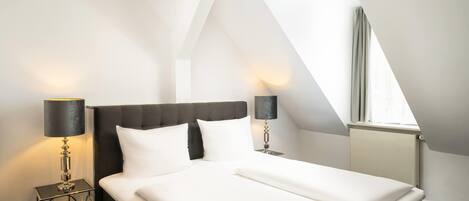 A modern hotel room with a large bed, two bedside tables with lamps, a window with curtains, and a sloped ceiling.