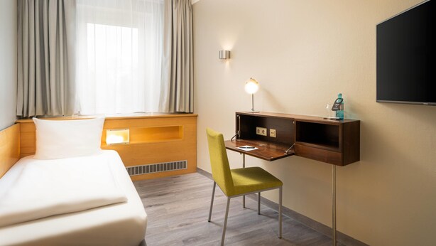 Une chambre d’hôtel comprenant un lit, un bureau avec une chaise, un téléviseur fixé au mur et une fenêtre avec des rideaux.