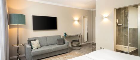 A modern hotel room with a bed, a sofa, a desk, a flat-screen TV, and a shower area.
