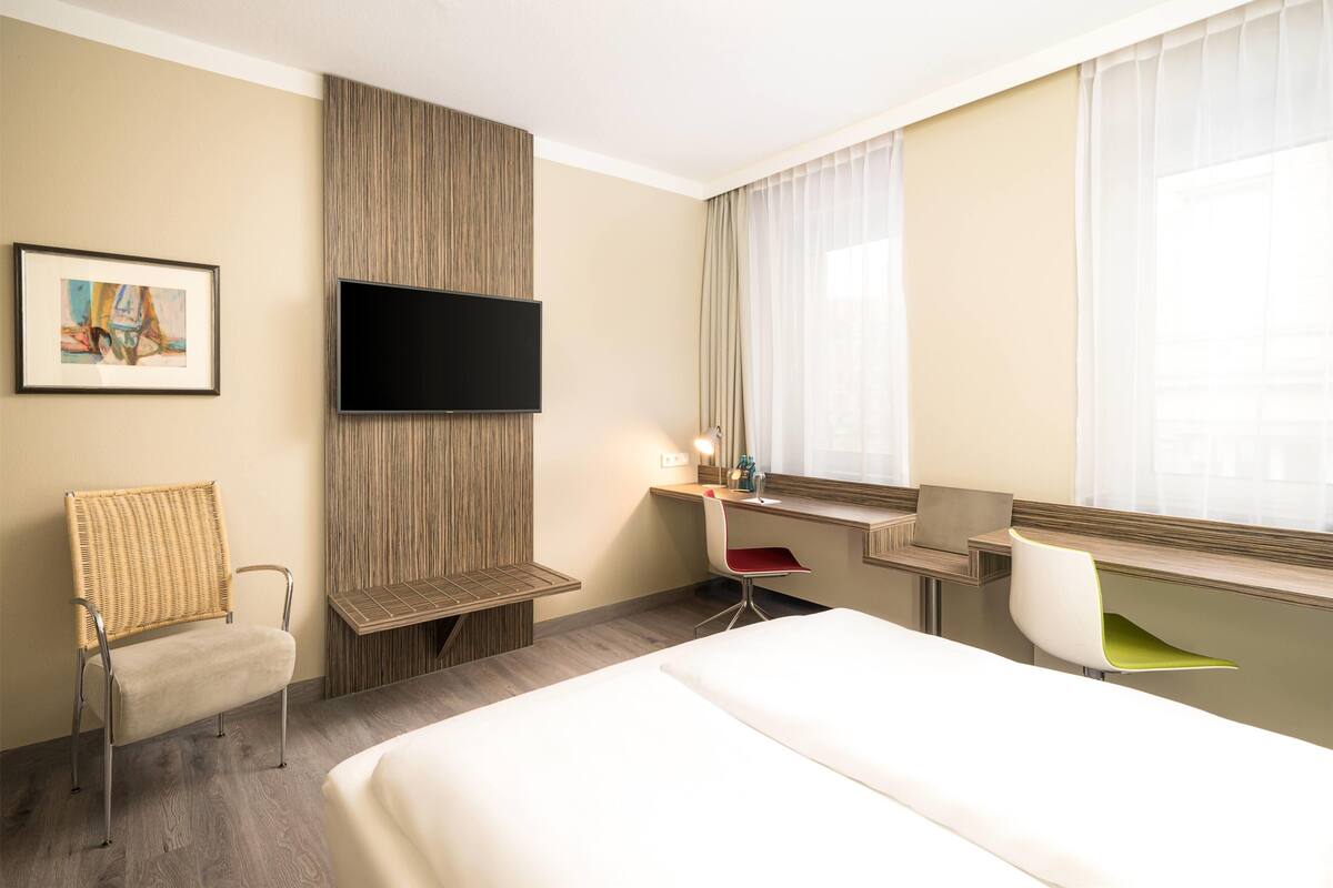 Una habitación de hotel con cama, escritorio, silla, televisor y ventana con cortinas.