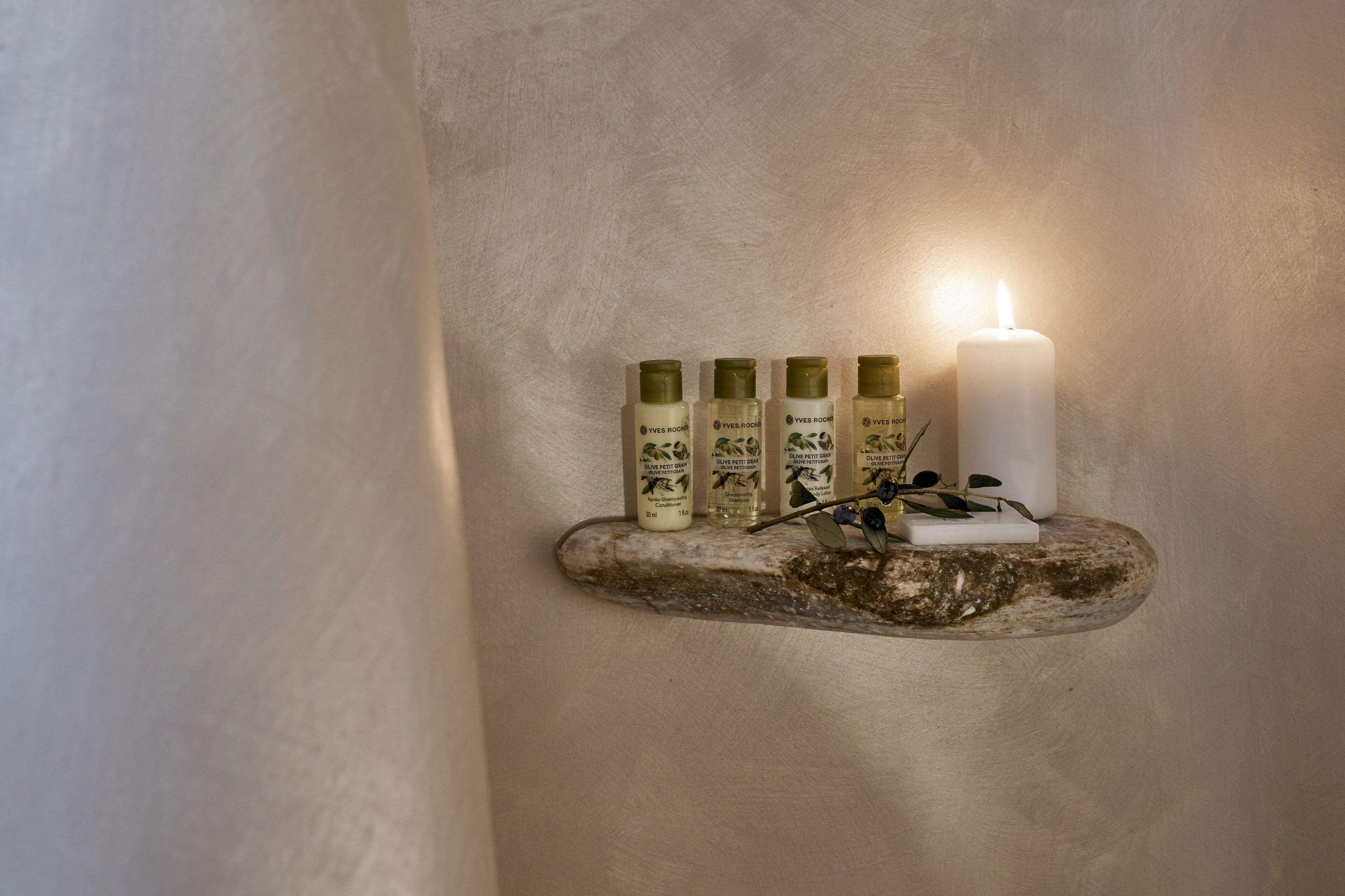 A shelf with four bottles and a lit candle against a textured wall.