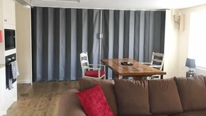 A living room with a brown sofa, a wooden dining table with chairs, a kitchen area with a microwave, and a large window with curtains. - No1 Dove Street (Ipswich)