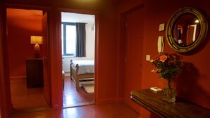 A hotel hallway with a wooden floor, a bed in the adjacent room, a mirror on the wall, and a vase with flowers on a table. - No1 Dove Street (Ipswich)