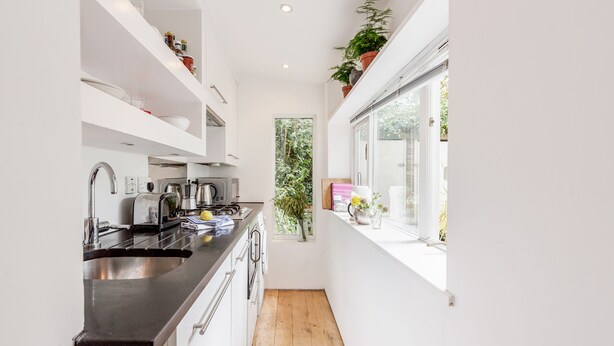 A narrow kitchen with white cabinets, a black countertop, and a window with a view of greenery. - Modern Holland Park apartment (London)