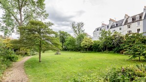 A grassy courtyard with a gravel path, surrounded by trees and a building with multiple windows and chimneys. - Modern Holland Park apartment (London)