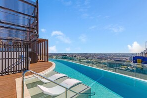 Une piscine à débordement située sur le toit, avec une eau d’un bleu cristallin, des chaises longues blanches et une vue sur la ville.