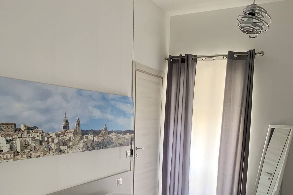 A hotel room with a bed, a cityscape painting, a mirror, and a window with curtains.