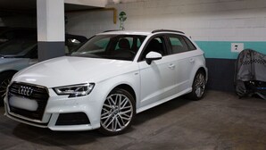 Un Audi station wagon blanco estacionado en un estacionamiento con pilares de concreto y piso de baldosas.