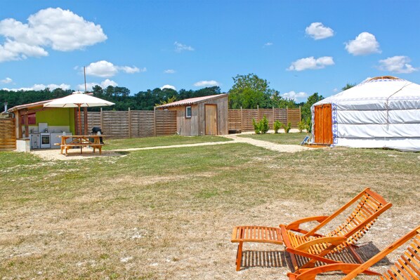 Une cour arrière clôturée avec une structure en bois, une tente blanche et deux chaises longues orange.