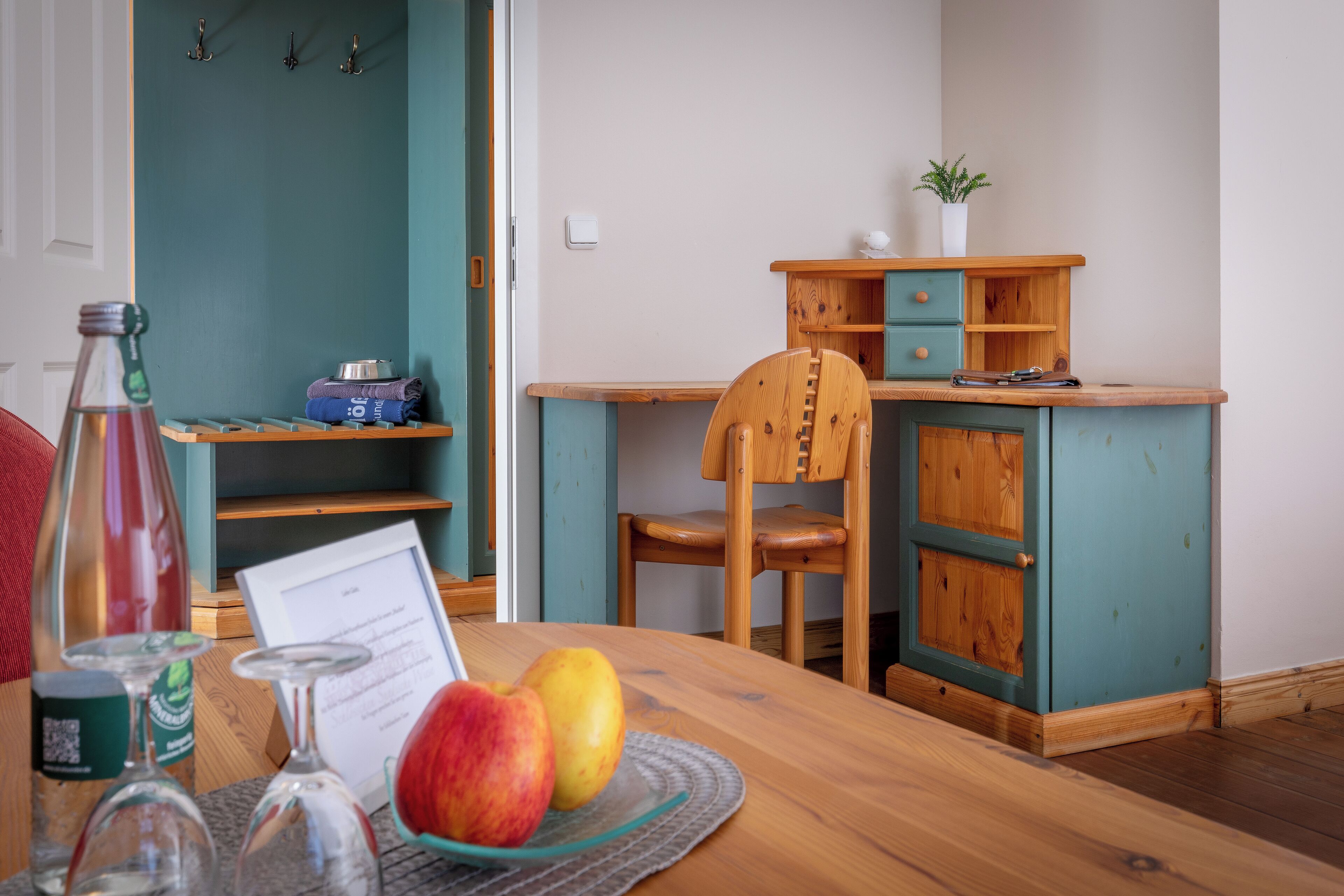 A wooden table with a glass plate holding two apples, a bottle, and a menu. A wooden chair is positioned at the table, and a teal-colored cabinet with a plant on top is visible in the background.