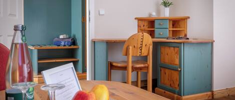 A wooden table with a glass plate holding two apples, a bottle, and a menu. A wooden chair is positioned at the table, and a teal-colored cabinet with a plant on top is visible in the background.