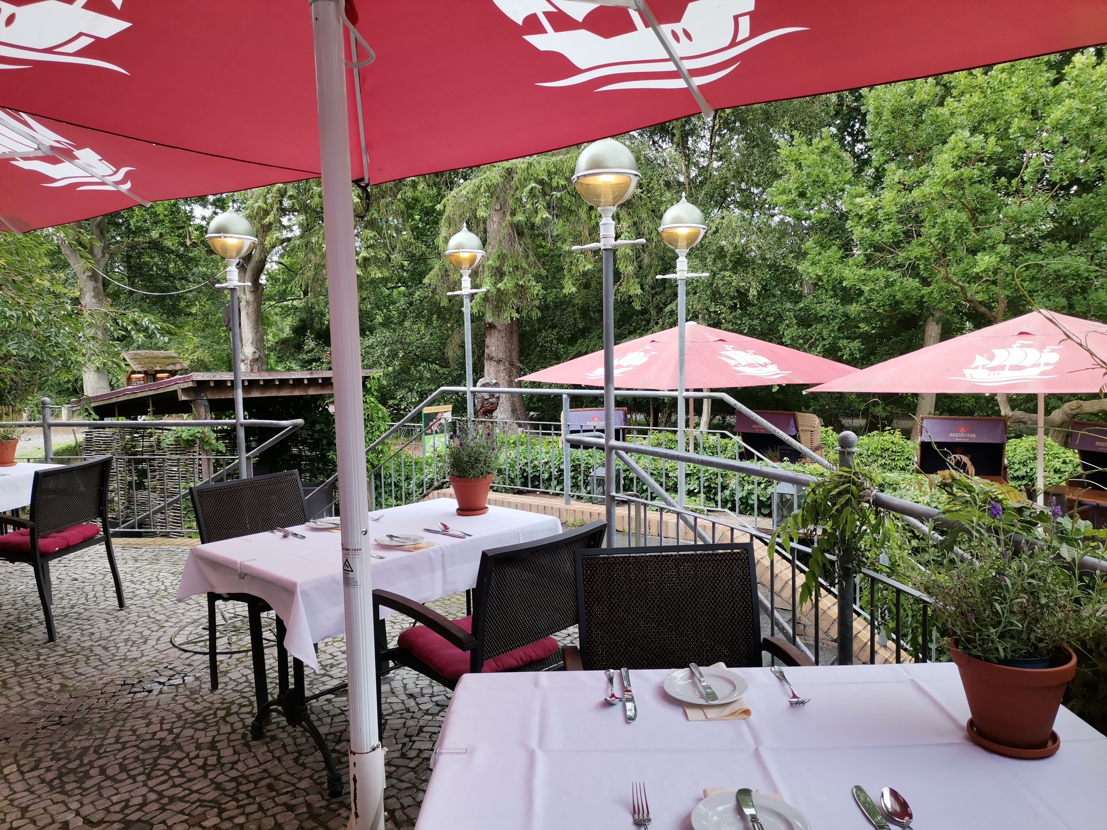 An outdoor dining area with tables and chairs under red umbrellas, surrounded by greenery.