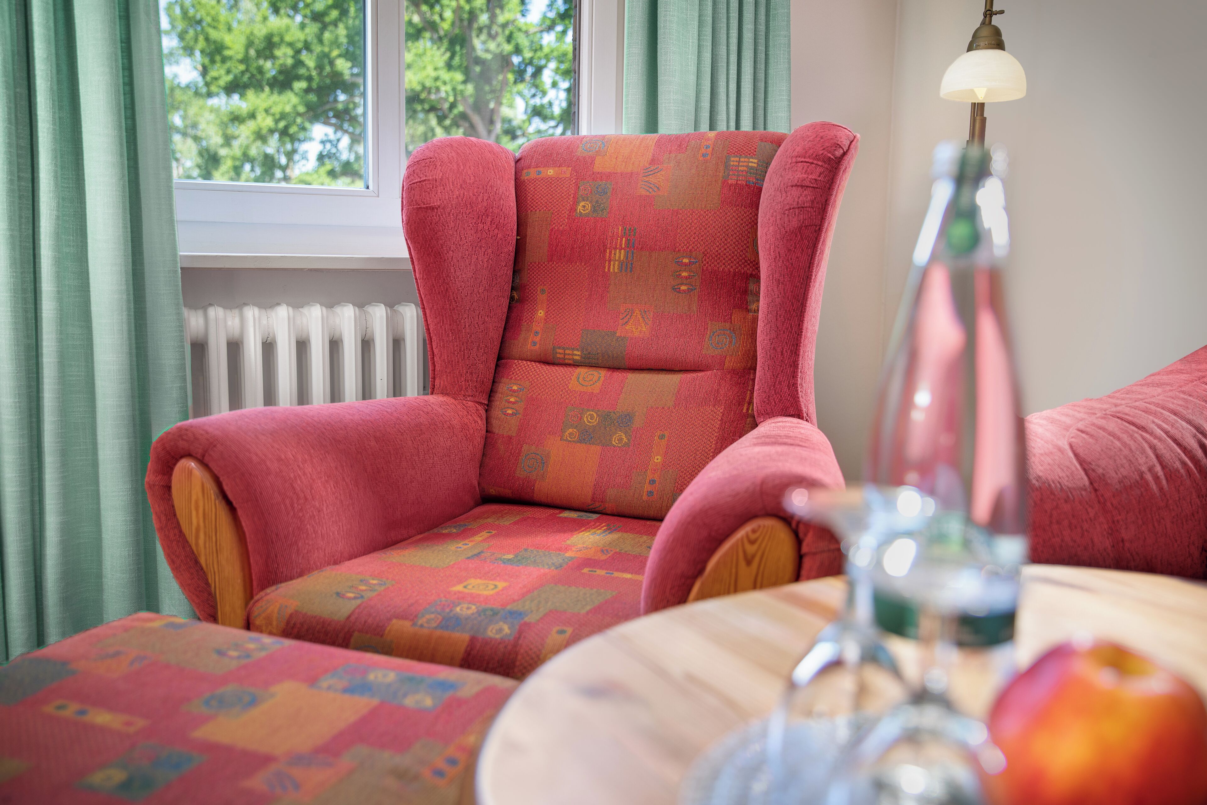 A cozy room with a patterned armchair, a table with a glass, and a window with curtains.