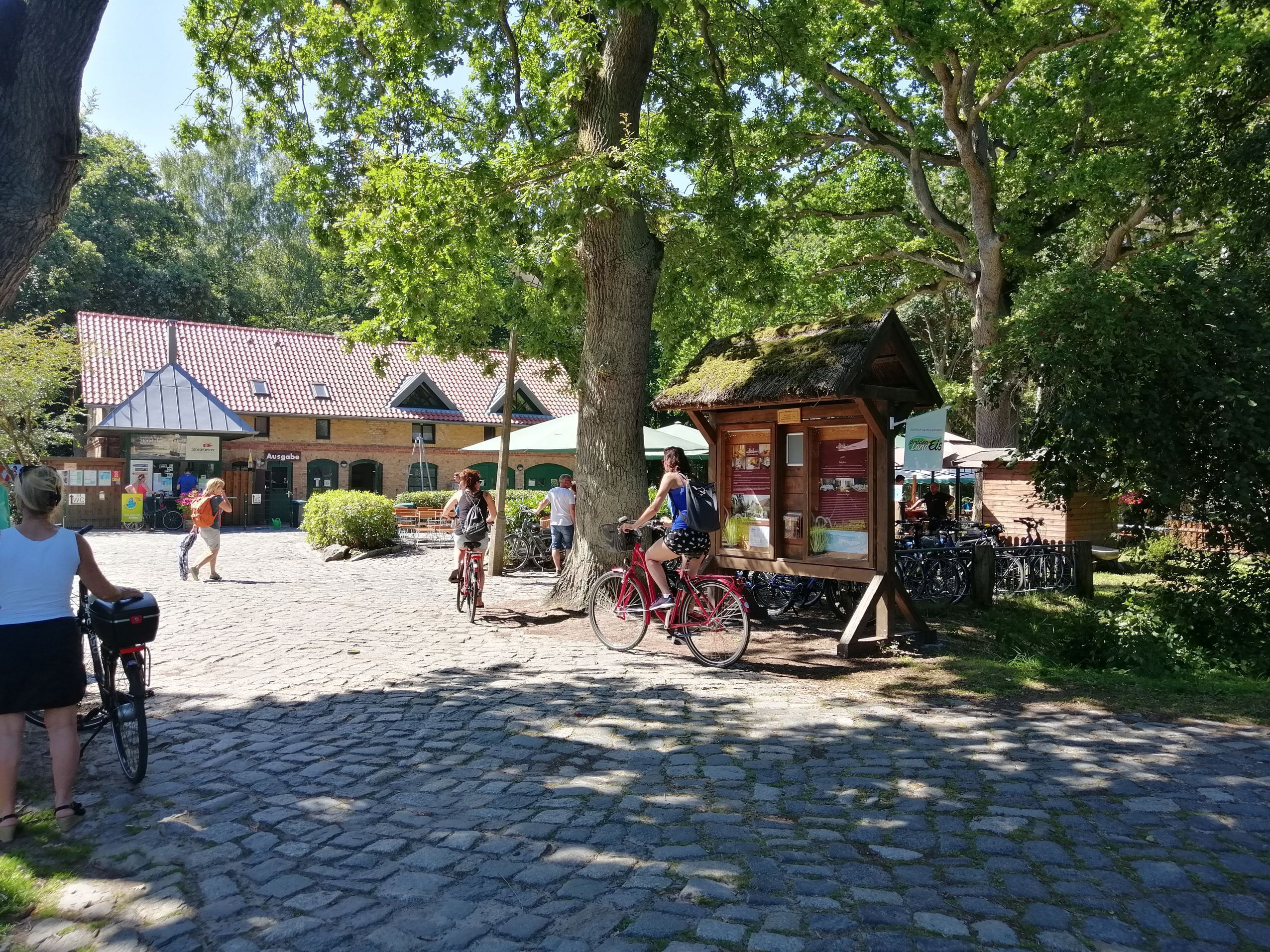 Un sentier pavé de pierres, des vélos, une structure en bois avec une enseigne et, au fond, un bâtiment avec un toit recouvert de tuiles rouges.