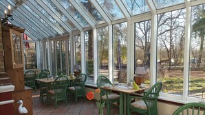Une véranda avec un toit en verre, des chaises vertes, une table en bois et une vue sur les arbres et un jardin.