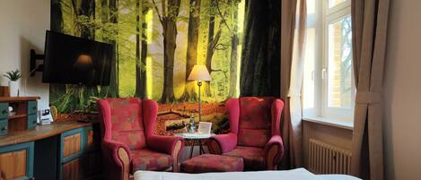 A hotel room with a large bed, two red armchairs, a small table, a TV, and a forest mural on the wall.