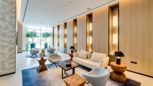 Um lobby de hotel moderno com um sofá branco, várias cadeiras, uma mesa de centro e uma janela grande com luz natural.