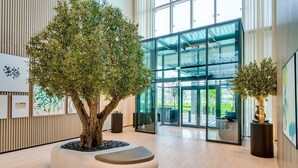 Um lobby moderno com um grande vaso de plantas com árvore, portas de vidro que dão para uma área externa e obras de arte emolduradas nas paredes.
