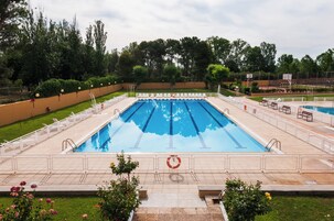 Una gran piscina al aire libre con varias pistas, rodeada por una valla, y una cancha de baloncesto al fondo.