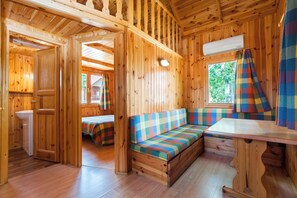 A cabin interior with a wooden floor, a bench with a checkered cushion, a table, and a window with a plaid curtain. - Bugalows camping Regio (Santa Marta de Tormes)