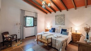 A bedroom with a large bed, two bedside tables, a bench, a chair, a window with curtains, and a decorative wall piece. - El Encinar Antequera - La Joya by Ruralidays (Málaga)