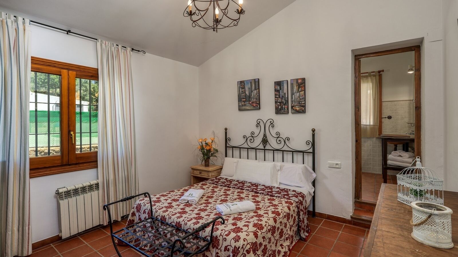 A bedroom with a bed, a nightstand, a window with curtains, a radiator, and a mirror in the adjacent room.