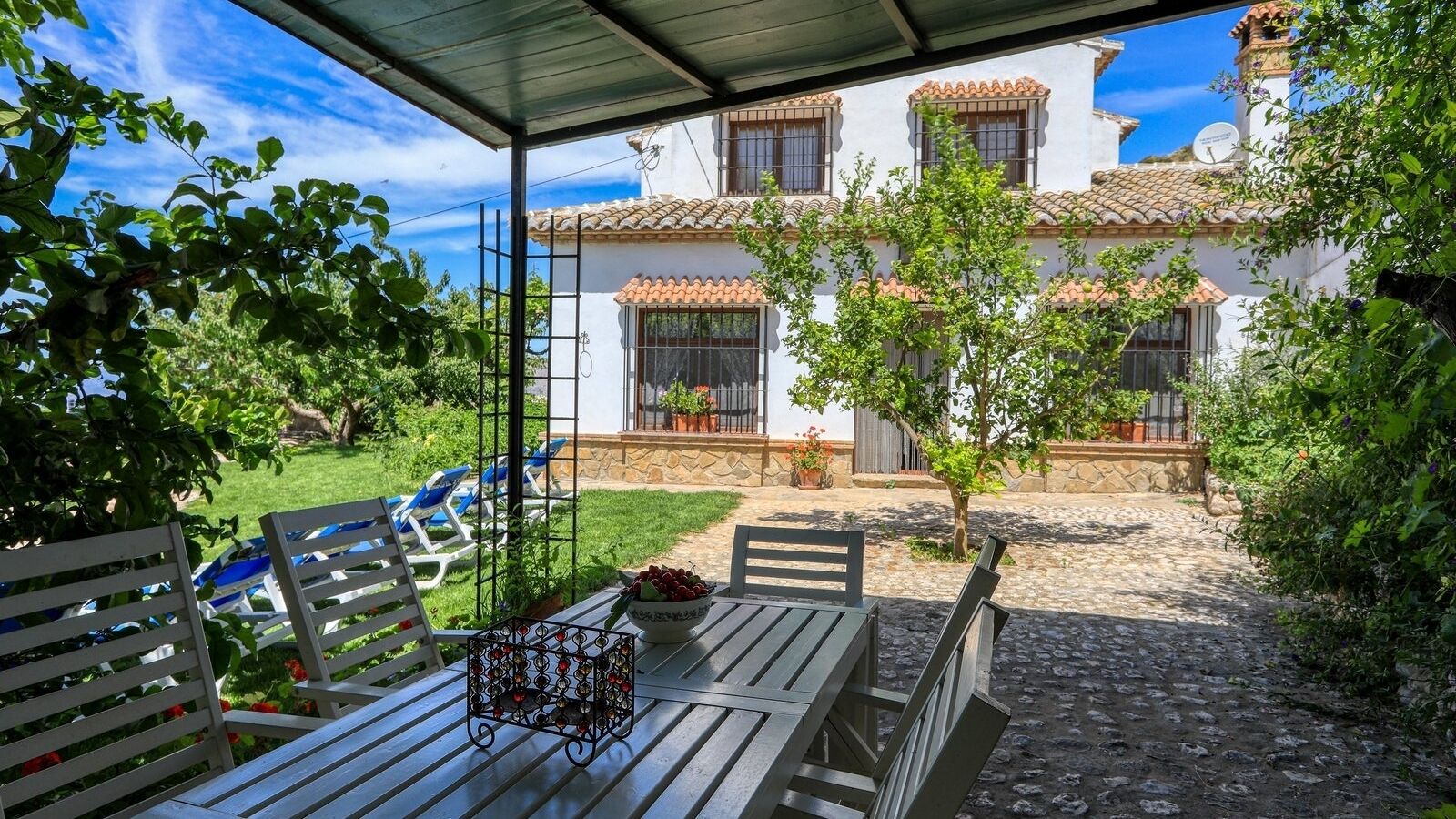 A covered patio with a table and chairs, a small garden, and a white building with red-tiled roofs in the background.