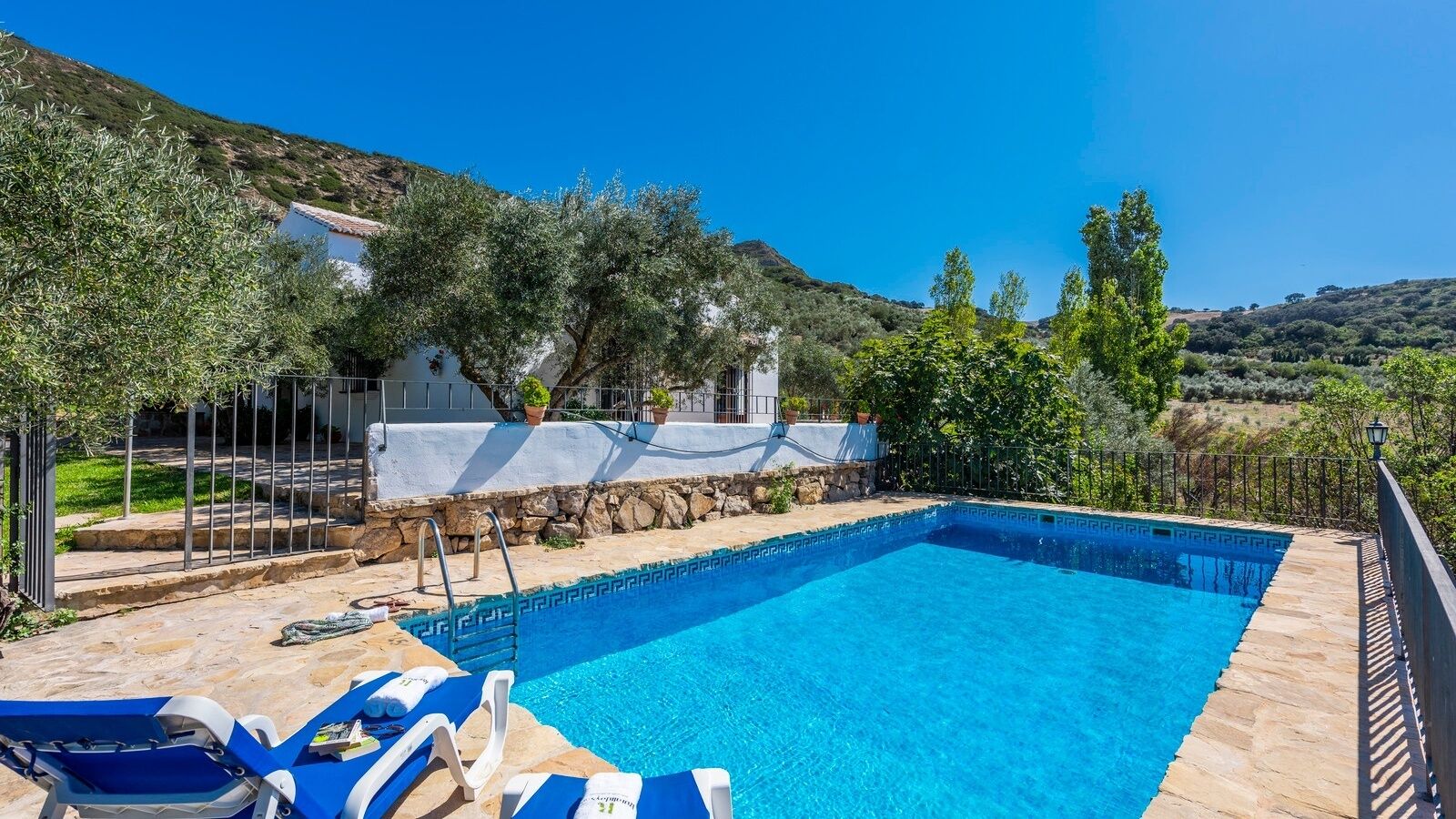 Una piscina rectangular con borde de piedra, rodeada por una terraza de piedra. Hay sillas de descanso y una pequeña mesa con una toalla. La piscina está junto a un edificio con fachada blanca y un muro de piedra.