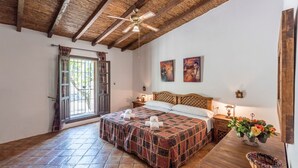 Une chambre avec un plafond en bois, un lit recouvert d’une couvre-lit à motifs, une table de chevet avec une lampe et une fenêtre avec des rideaux.