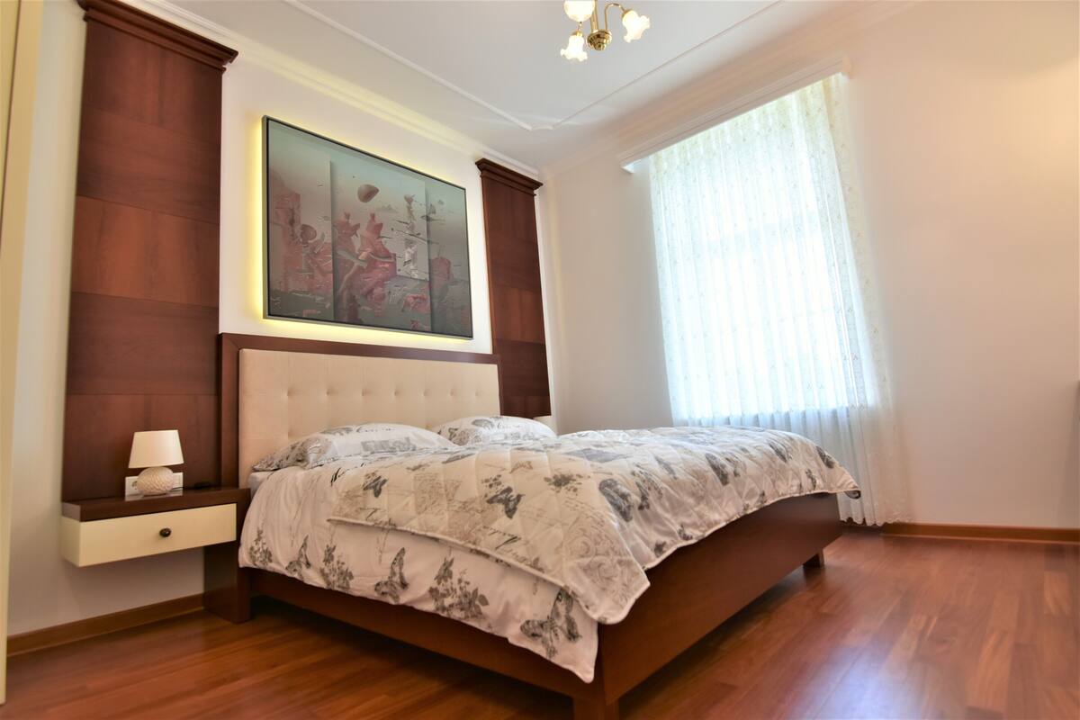 Een slaapkamer met een groot bed, een nachtkastje, een schilderij boven het bed en een raam met gordijnen.