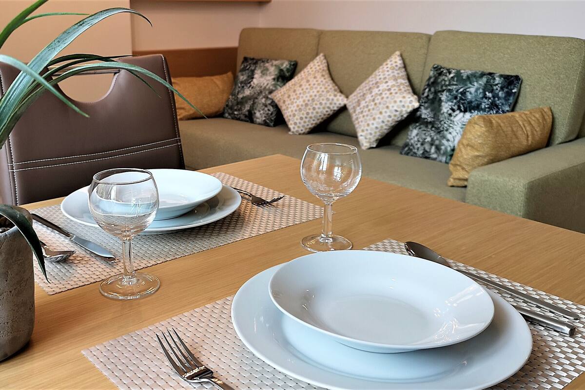 Een eettafel gedekt met borden, glazen en bestek, een potplant en een bank met gekleurde kussens op de achtergrond.