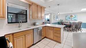A modern kitchen with wooden cabinets, a dishwasher, and a dining area with a table and chairs. - Shady Oaks - Breezy retreat w patio & beach access (Jacksonville Beach)