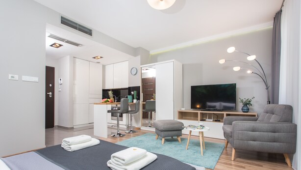 A modern hotel room with a bed, a TV, a grey armchair, a small table, and a kitchenette area. - Studio Apartment, 15 min. walk to Main Square, 10 min. walk to Jewish Quarter (Grzegórzki)