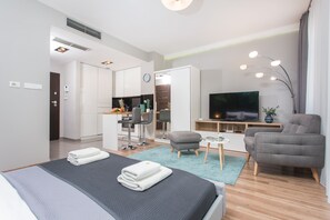 Quarto de hotel moderno com cama, televisão, poltrona cinza, mesa pequena e área de cozinha compacta.