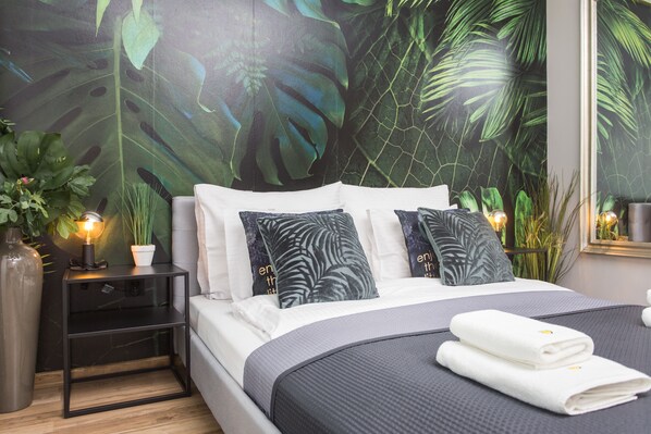 Quarto com uma cama grande, duas mesas de cabeceira, um vaso de planta e um mural na parede.
