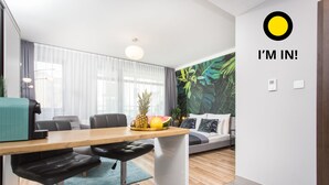 A modern living room with a wooden dining table, two black bar stools, a sofa with patterned cushions, a large window with sheer curtains, and a tropical-themed wall mural. - Studio Apartment, 15 min. walk to Main Square, 10 min. walk to Jewish Quarter (Grzegórzki)